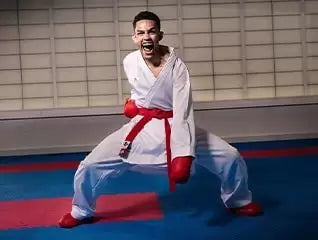 Karate athlete in a white gi with a red belt and gloves performing a powerful stance and shout on a dojo mat.