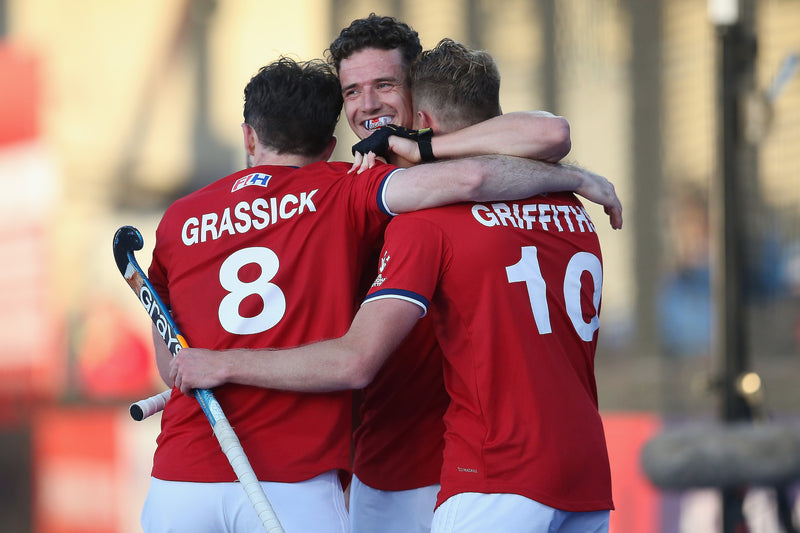 England men’s hockey players celebrating a goal, wearing red kits with names Grassick and Griffiths visible, embracing on the pitch.
