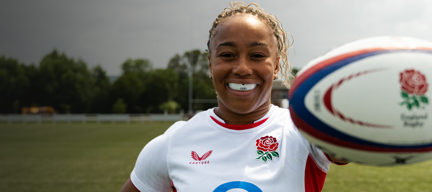 Sadia Kabeya wearing a white Opro mouthguard in England rugby jersey holding a rugby ball on a field.