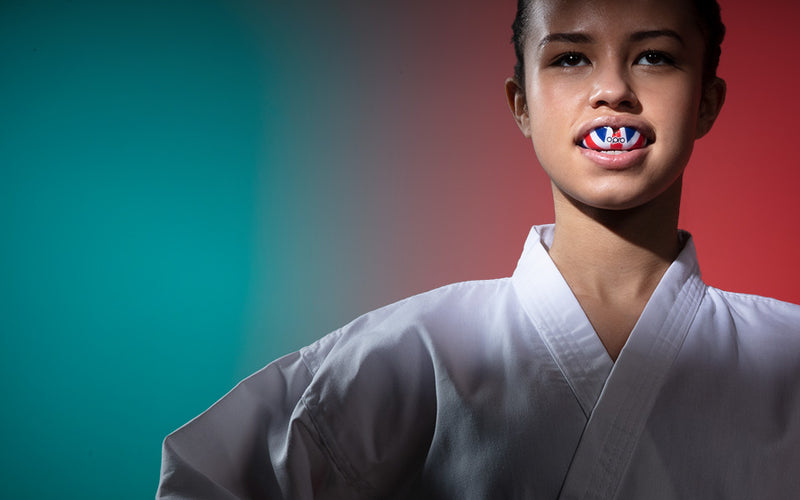 Young taekwondo athlete wearing a white uniform, confidently showing a red, white, and blue OPRO mouthguard, with a split teal and red background.