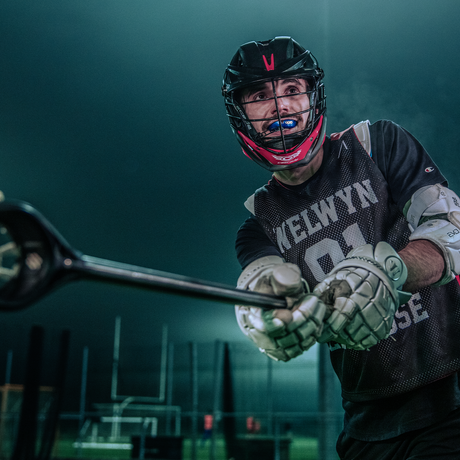 Lacrosse player wearing a helmet and protective gear holds a lacrosse stick, showing a blue OPRO mouthguard.