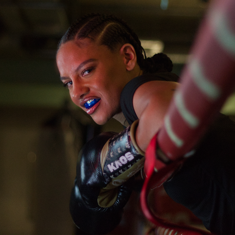 Female boxer wearing braided cornrows, black boxing gloves, and a blue Opro mouthguard leans against the ropes in a boxing ring, looking determined and confident.