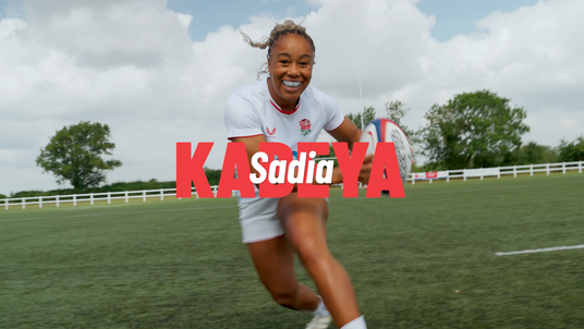 England rugby player Sadia Kabeya runs with a rugby ball on the pitch, wearing the England kit and smiling, with her name “Sadia Kabeya” in bold red and white text across the image.
