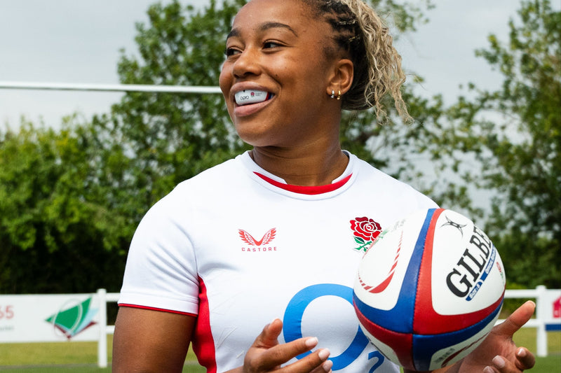 Smiling female rugby player in an England kit holding a Gilbert rugby ball, wearing a white OPRO mouthguard on the pitch during training.