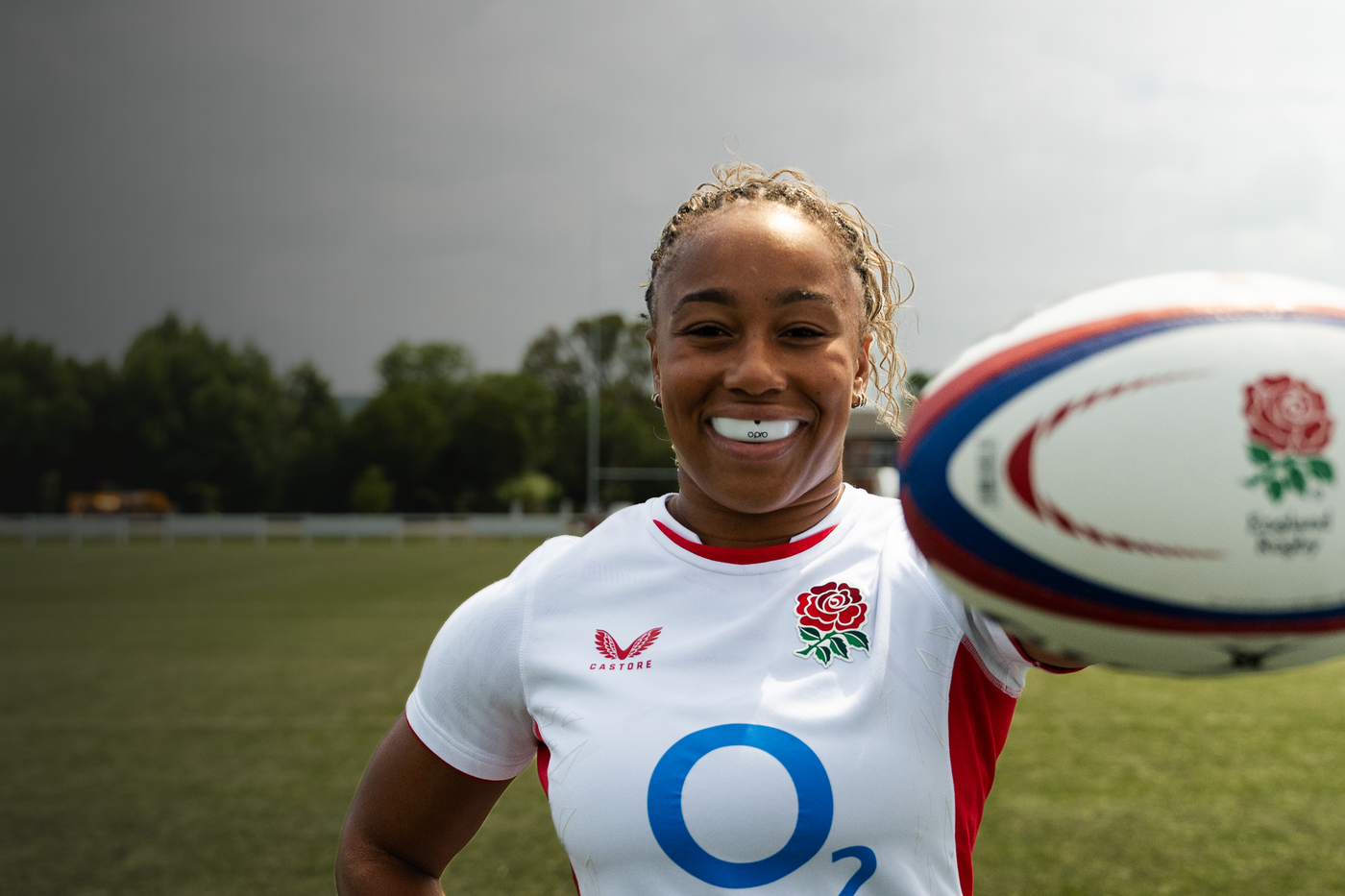Sadia Kabeya wearing a white Opro mouthguard in England rugby jersey holding a rugby ball on a field.