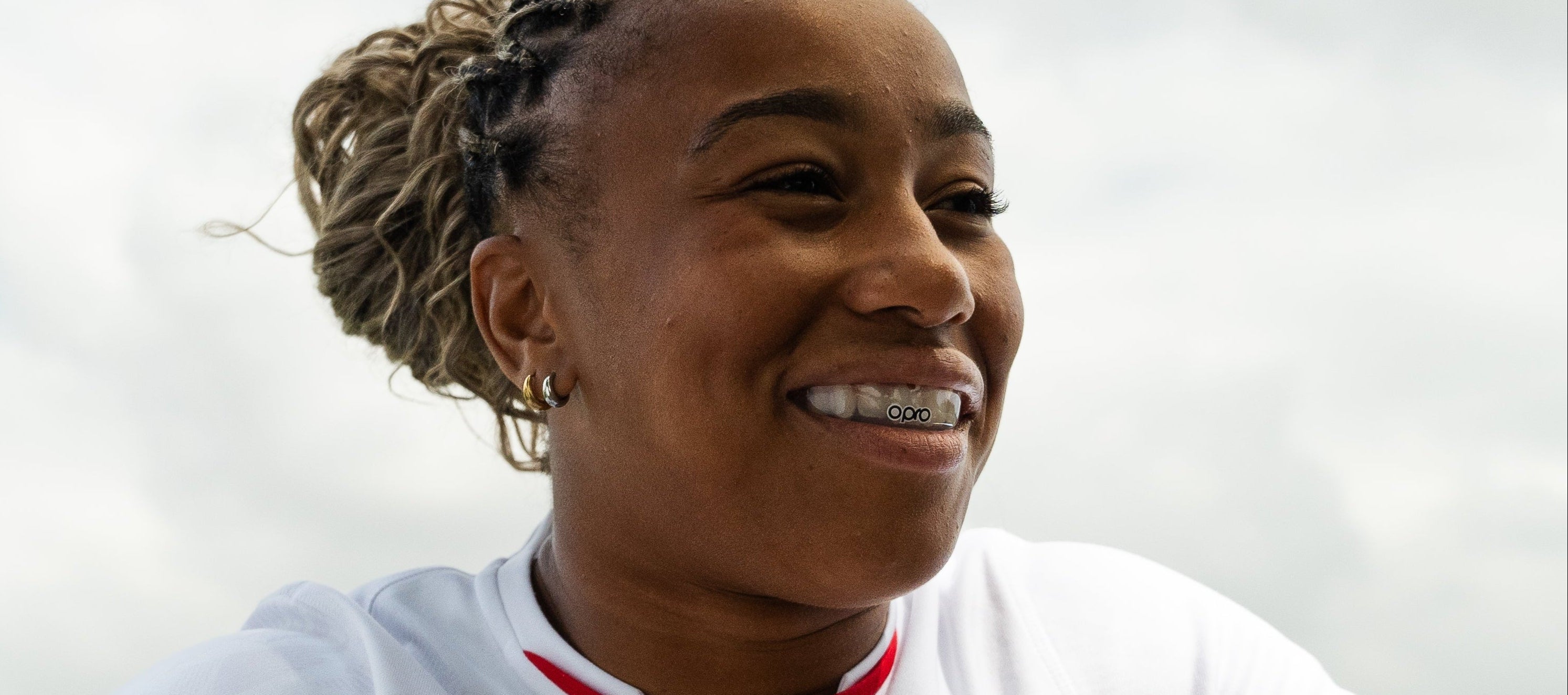 Smiling women’s rugby player wearing an England jersey holds a rugby ball and shows her OPRO clear mouthguard on the pitch.