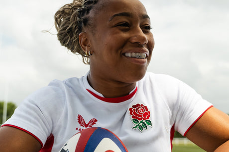 Smiling women’s rugby player wearing an England jersey holds a rugby ball and shows her OPRO clear mouthguard on the pitch.
