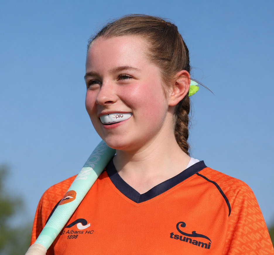 A smiling young woman outdoors wearing an orange athletic jersey with navy trim and holding a light blue and white bat. The jersey has a visible 'tsunami' logo on the chest.
