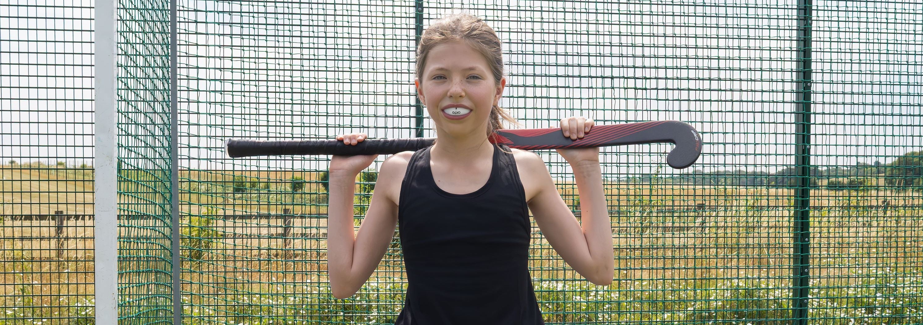 A young girl with blonde hair stands on a blue court inside a netted practice facility, holding a field hockey stick over her shoulders. She is wearing a black tank top and a white skirt.