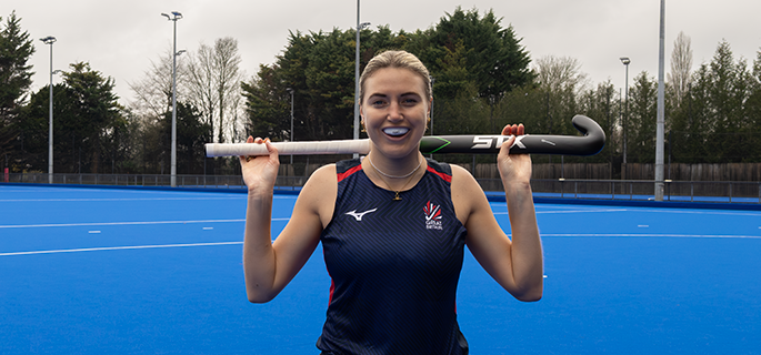 A woman stands on a blue field hockey pitch holding a hockey stick over her shoulders, wearing a dark navy sleeveless athletic top and matching skirt with the number 26 on the front.