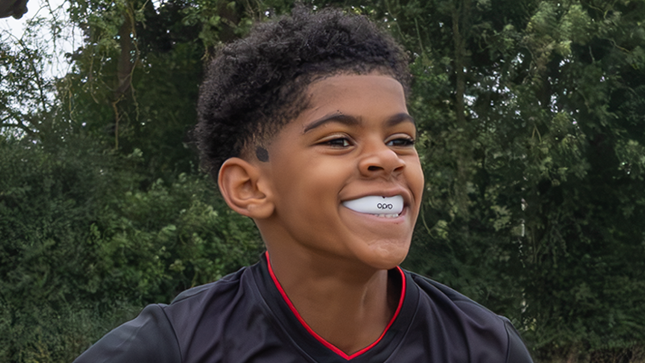 A young boy smiling outdoors while wearing a black rugby jersey with red trim and holding a white rugby ball with red and blue stripes. Trees and grass are visible in the background.