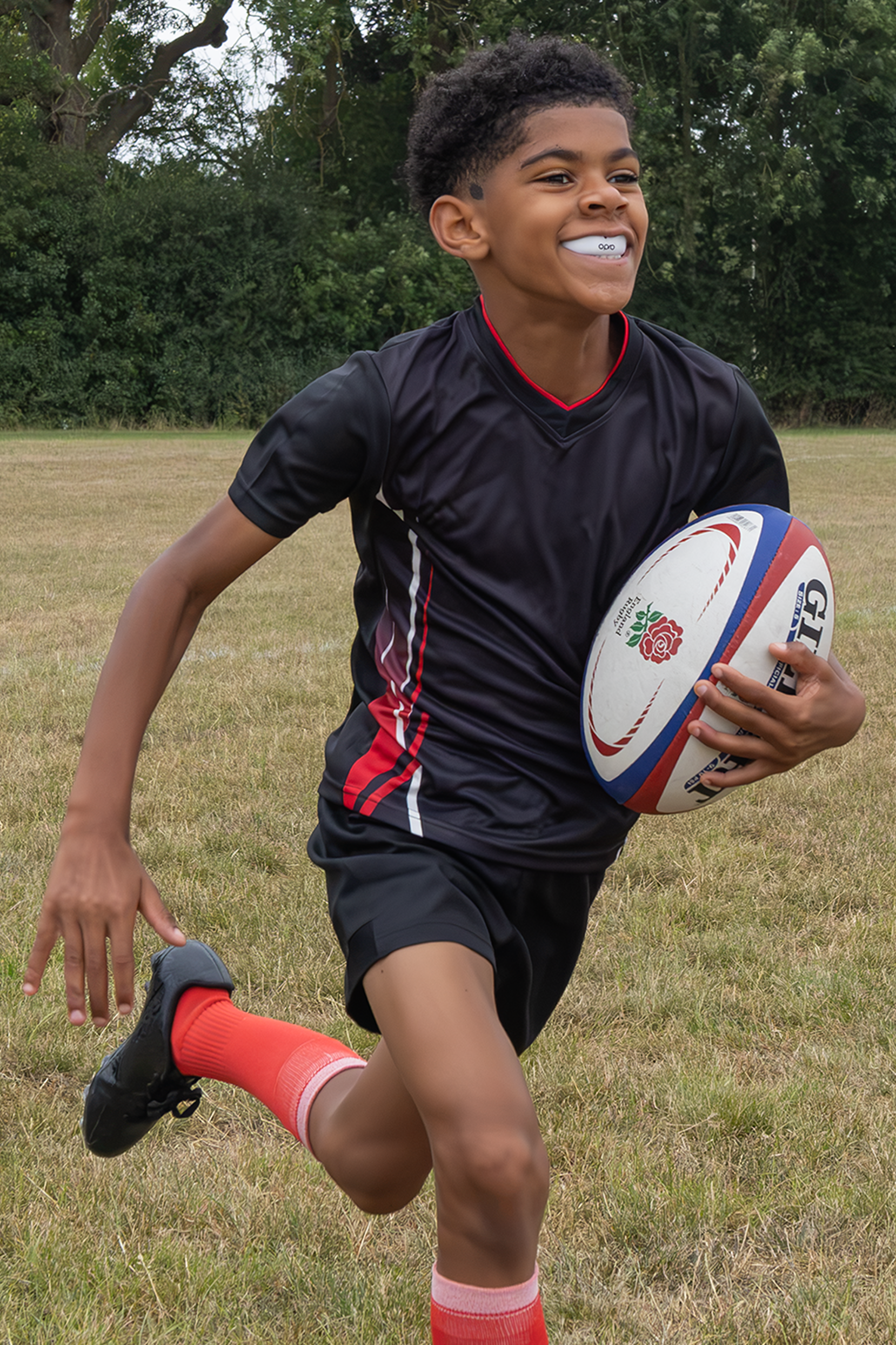 A young boy wearing a black rugby jersey and shorts with red accents, running on a grassy field while holding a white rugby ball with red and blue stripes.