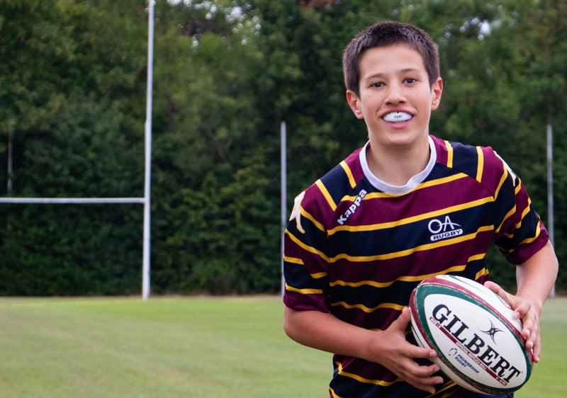 Young boy holding a rugby ball on a grassy field with trees in the background