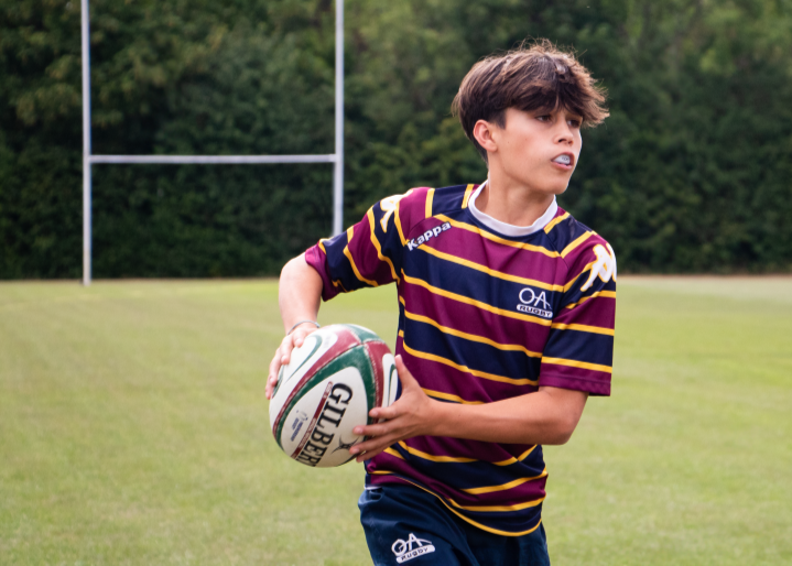 Young boy holding a rugby ball on a grassy field with trees in the background