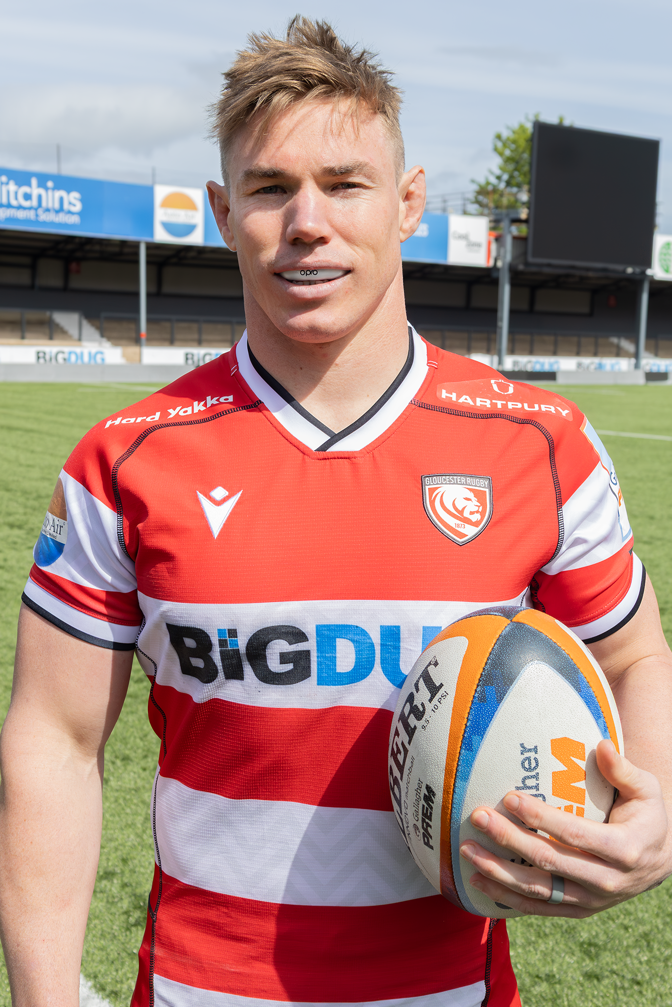 A male rugby player stands on a grass field holding a rugby ball, wearing a red and white striped rugby jersey with 'BIGDUCK' on the front, black shorts, and a rugby ball under his arm.