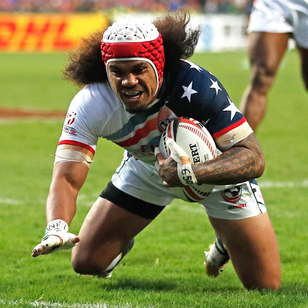 A rugby player in a red, white, and navy USA team jersey dives on a green field while holding a rugby ball. Another player is blurred in the background.