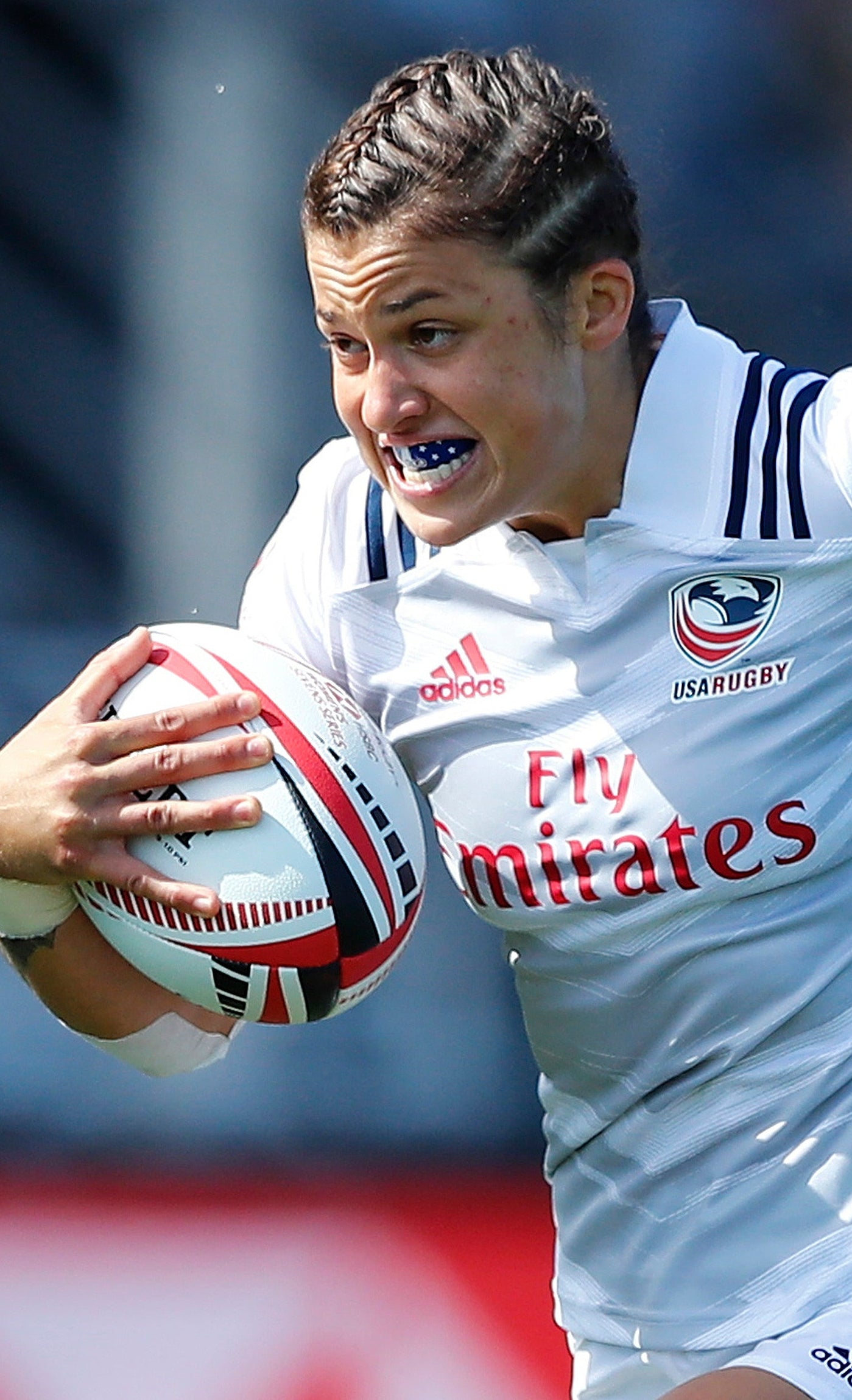 A rugby player in a white Southwestern Warriors jersey with red and black accents runs while holding a rugby ball, with another player blurred in the background.