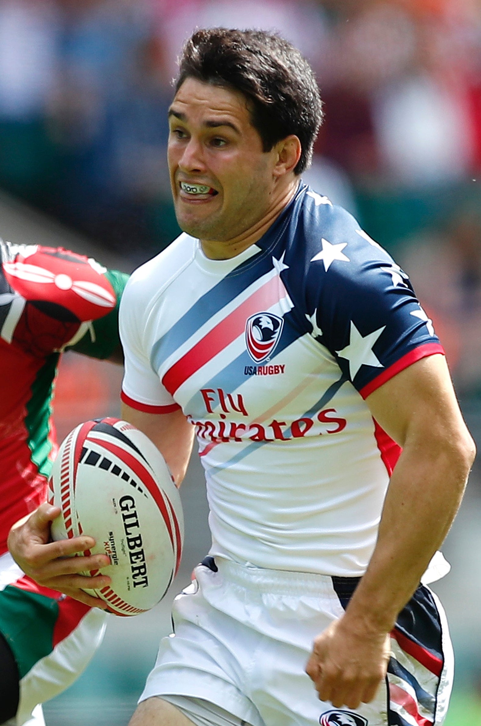 A rugby player in a white, red, and blue jersey carrying a Gilbert rugby ball, being chased by an opponent in a red jersey on a stadium field.