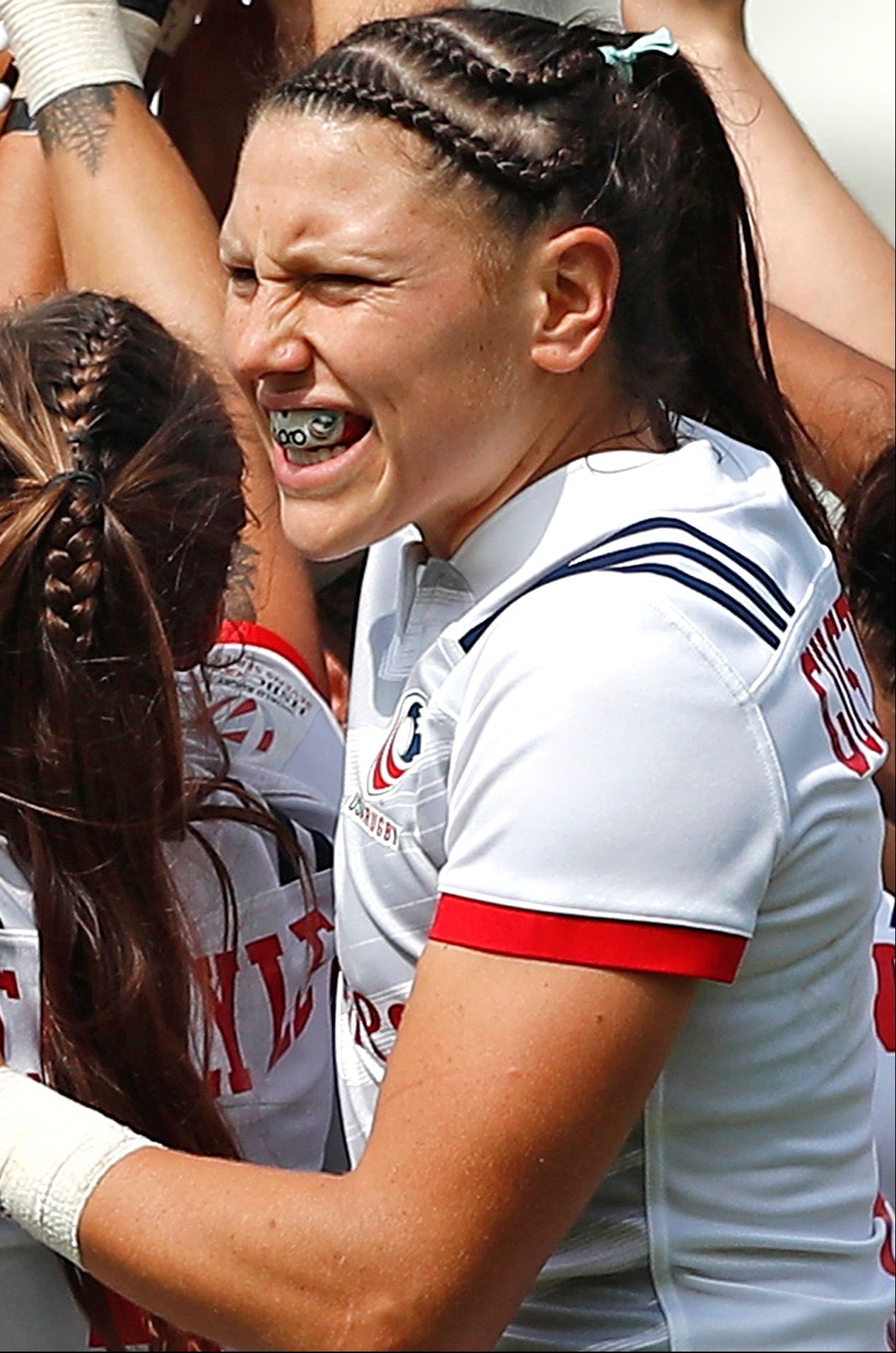 A group of female rugby players in white jerseys with red and navy accents celebrate together, with one player in the center smiling broadly while others raise their hands in jubilation.