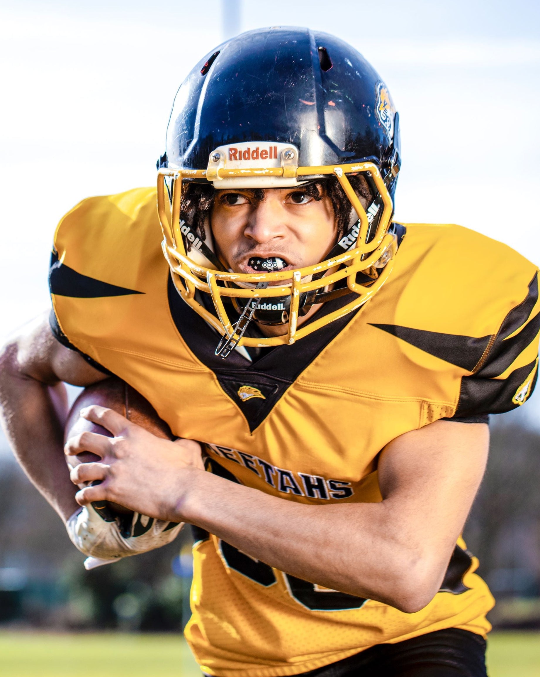 A football player in a yellow and black uniform with a black Riddell helmet carrying an American football on a field.
