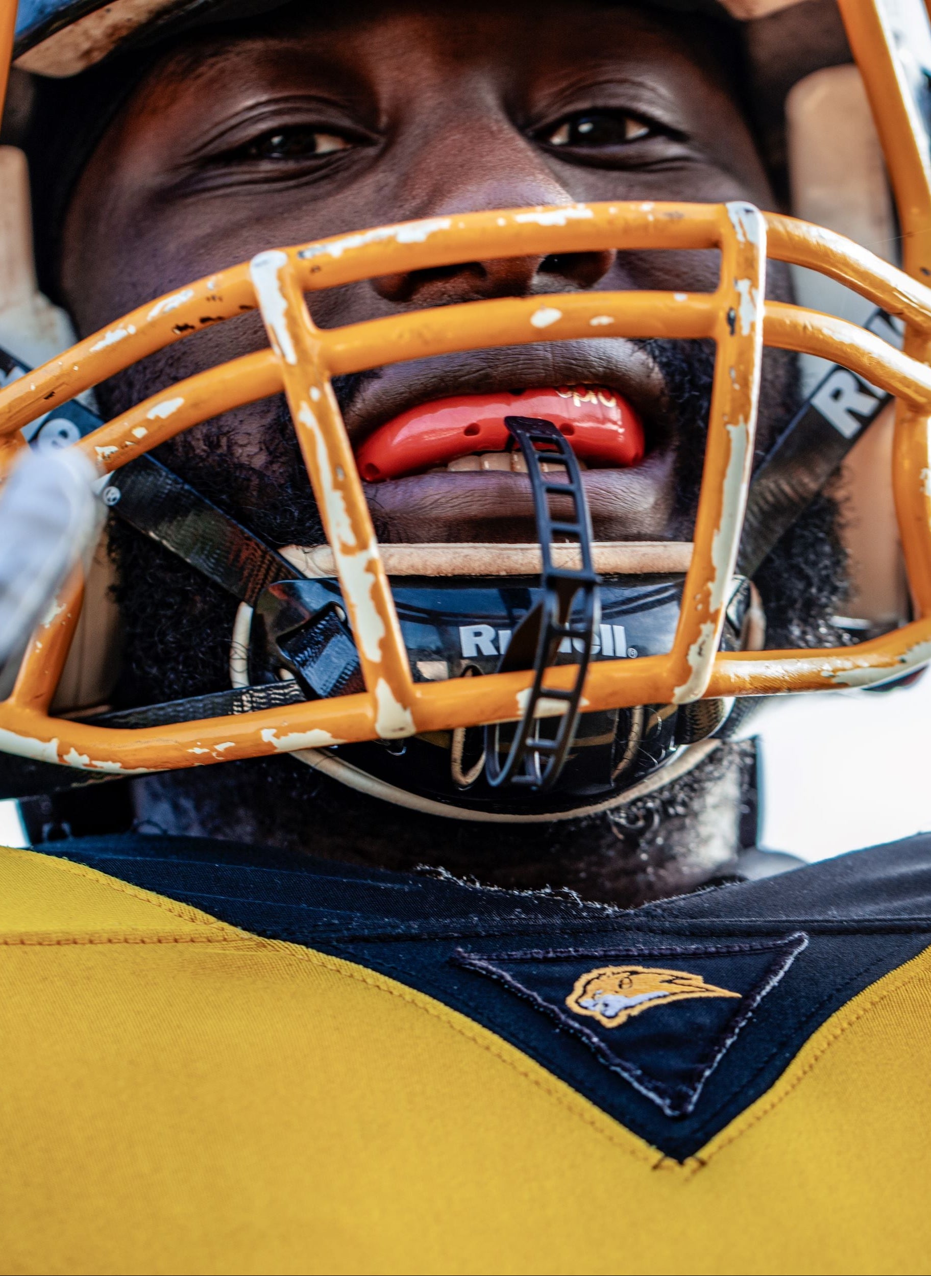 Close-up of an American football player wearing a yellow and navy uniform, a gold faceguard, a dark helmet, and white gloves, with a blurred outdoor sky background.