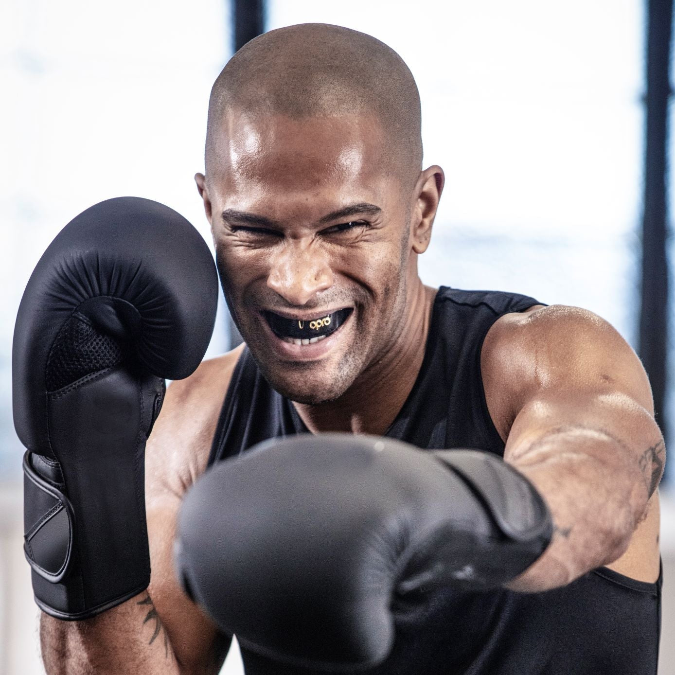 A smiling male boxer in a ring wearing black boxing gloves, a black tank top, and black shorts with a white waistband, captured mid-punch in a gym setting.