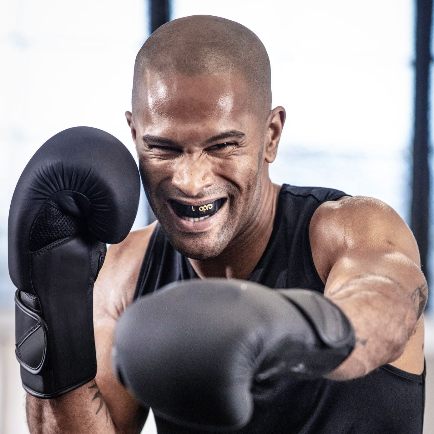 A smiling male boxer in a ring wearing black boxing gloves, a black tank top, and black shorts with a white waistband, captured mid-punch in a gym setting.