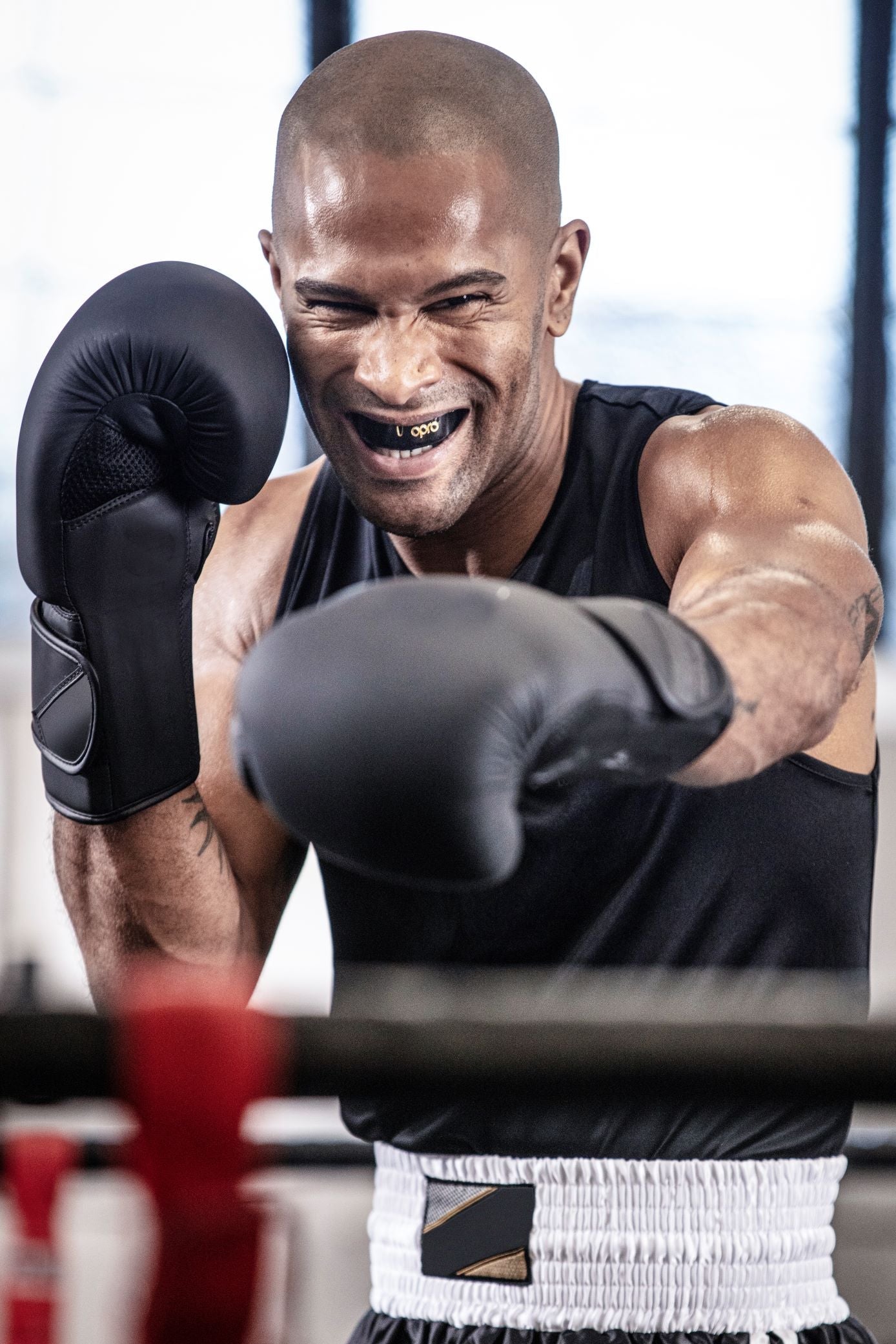 A smiling male boxer in a ring wearing black boxing gloves, a black tank top, and black shorts with a white waistband, captured mid-punch in a gym setting.