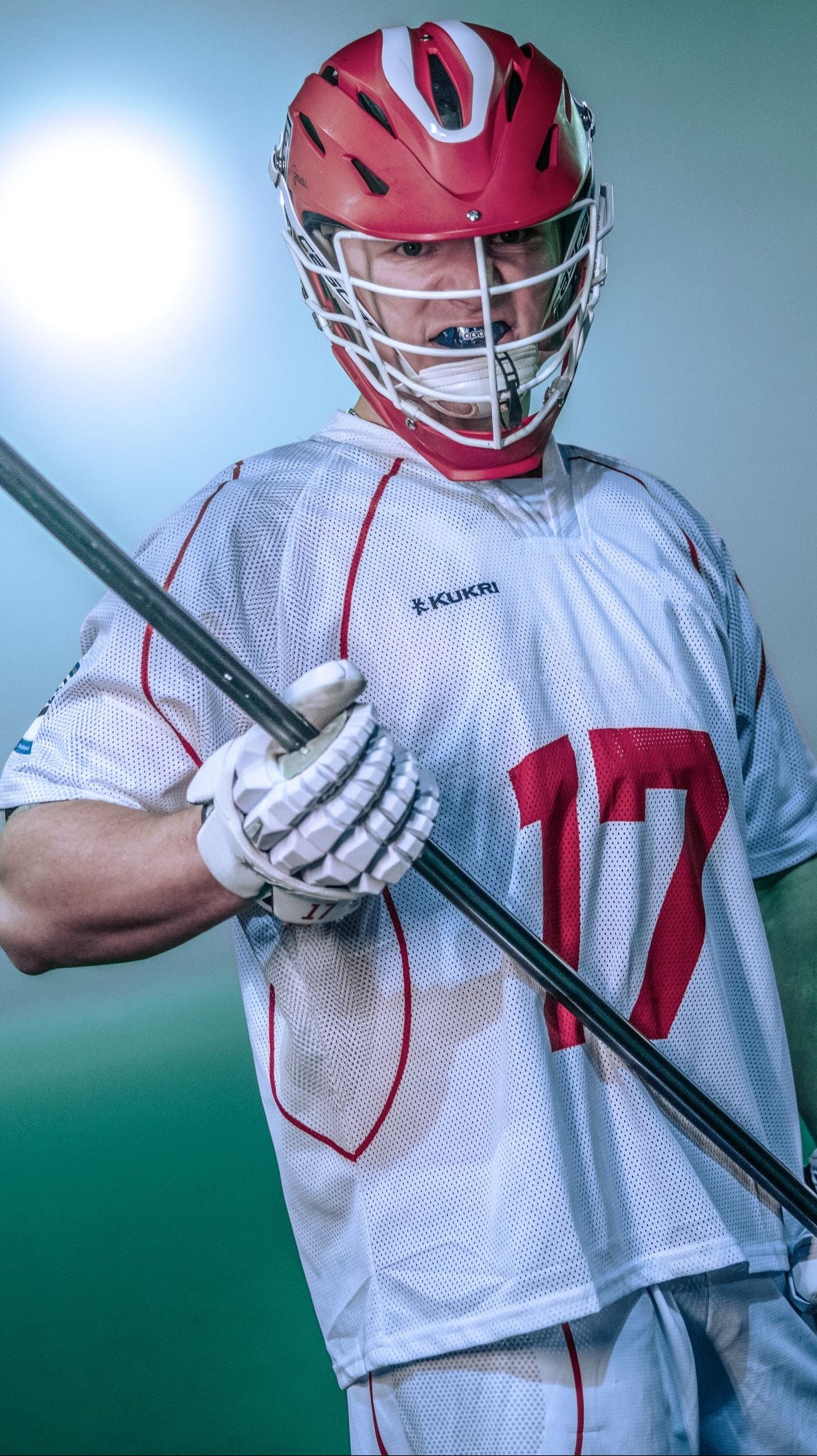 A lacrosse player wearing a red helmet, white jersey with red number 17, and holding a lacrosse stick, photographed against a moody green and gray background with a bright spotlight behind him.