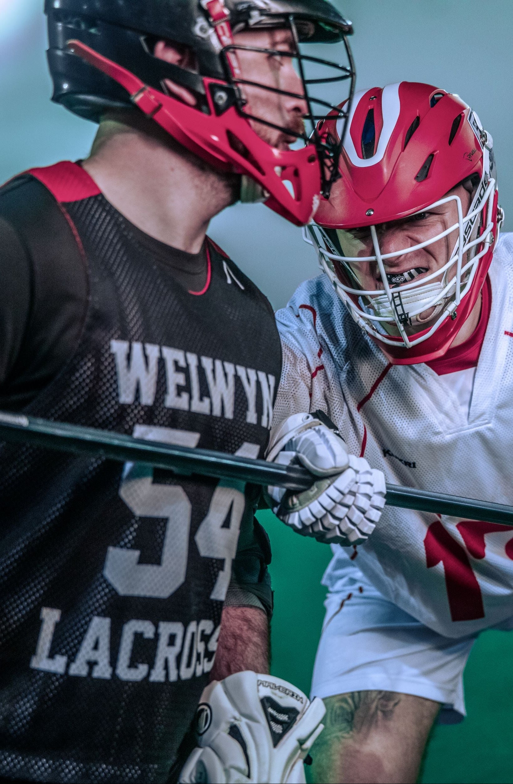 Two lacrosse players in action on an indoor turf field. One player wears a black and red jersey with 'WELVIVLACROS' and the number 54, while the other wears a white jersey with red numbers and a red helmet.