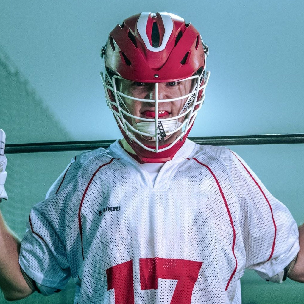 A lacrosse player in a red and white uniform holding a lacrosse stick in front of a net, with other players blurred in the background.