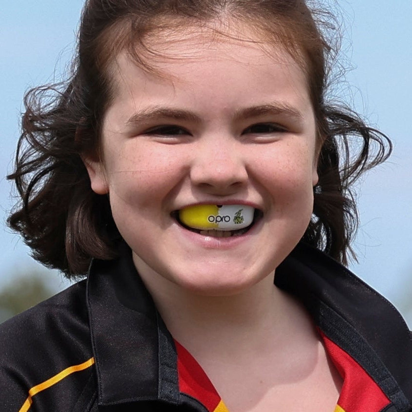 A close-up lifestyle shot of a smiling young woman with brown hair biting down on a yellow and white OPRO mouthguard, wearing a black and red sports jacket against a light blue sky background.