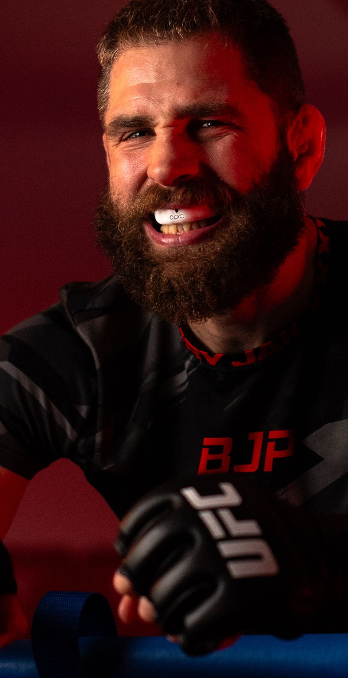 A close-up portrait of a bearded man in a black athletic shirt with red 'BJP' lettering and a black UFC glove, set against a dark red background.