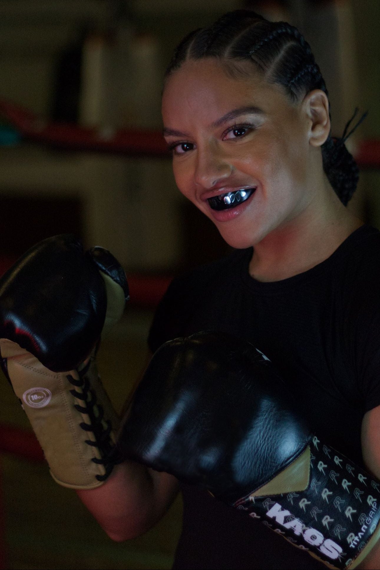 A woman in a boxing ring wearing black boxing gloves with KAOS branding, a black t-shirt, and a gold mouthguard, looking directly at the camera.